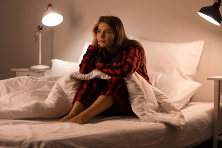 Upset young woman sitting on bed at nightの写真素材