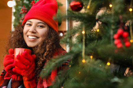 Beautiful young woman drinking hot tea near Christmas tree outdoors