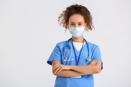 Female African-American doctor wearing medical mask on light backgroundの写真素材