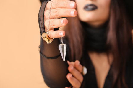 Scary young witch with pendulum on color background, closeup