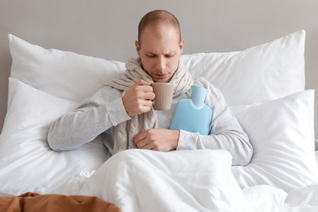 Ill man with cup of tea and hot water bottle in bedroomの写真素材