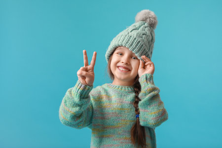 Stylish little girl in winter clothes showing victory gesture on color backgroundの写真素材
