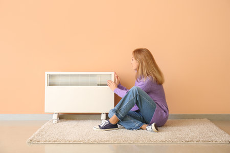 Mature woman near electric heater at home. Concept of heating seasonの写真素材