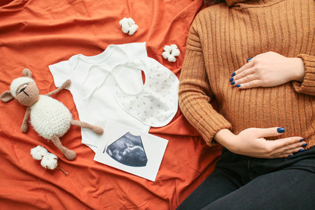 Pregnant woman with sonogram image and baby accessories on bedの写真素材
