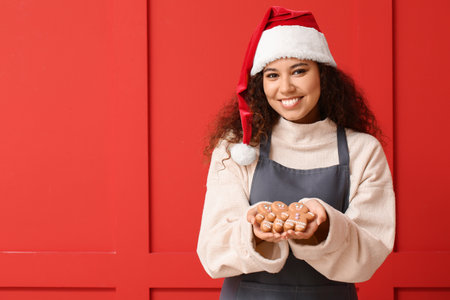 Beautiful young woman in Santa hat and tasty gingerbread cookies on color backgroundの写真素材