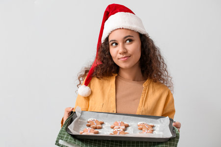 Beautiful young woman in Santa hat and tasty gingerbread cookies on light backgroundの写真素材