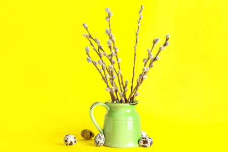 Jug with pussy willow branches and quail eggs on yellow background