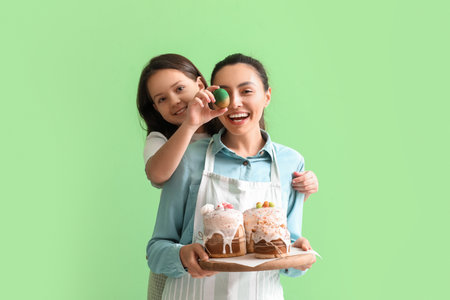 Beautiful woman and her little daughter with Easter egg and cakes on green background