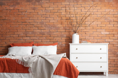 Vase with tree branches, books on chest of drawers and bed near brick wallの写真素材