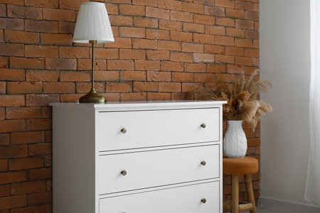 Chest of drawers with lamp, stool and vase near brick wall