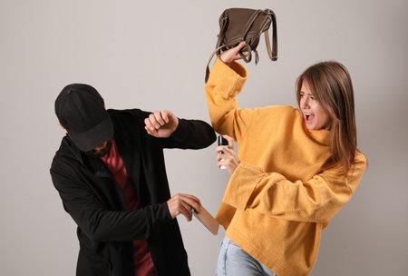 Young woman with pepper spray defending herself against thief on gray background
