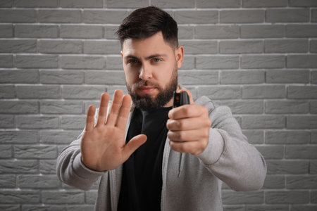 Young man with pepper spray for self-defence showing STOP gesture on gray brick background