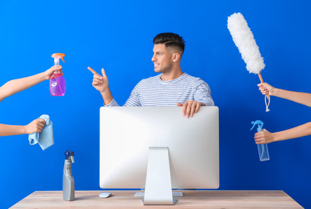 Young man, table with computer and hands holding different cleaning supplies on color backgroundの写真素材