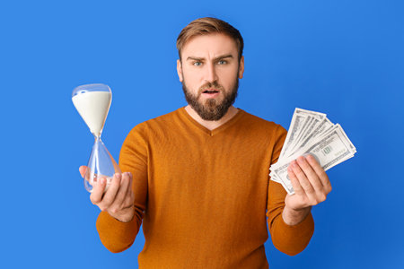 Young man with hourglass and money on color background