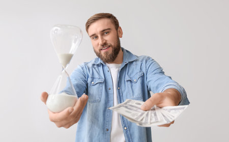 Young man with hourglass and money on light background