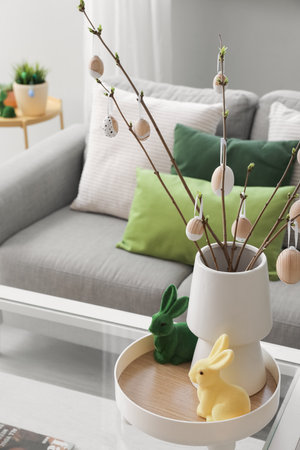 Vase with tree branches, Easter eggs and rabbits on table in light living room