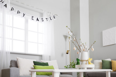 Vase with tree branches, Easter eggs and rabbits on table in light living room