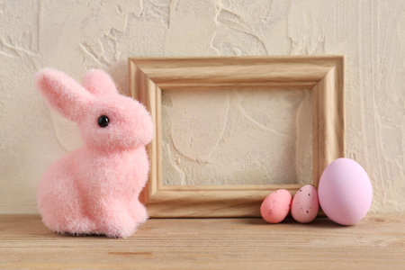 Empty picture frame, painted Easter eggs and bunny on wooden table near light wall