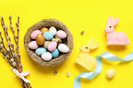 Composition with nest of Easter eggs, willow branches and toy bunnies on yellow background