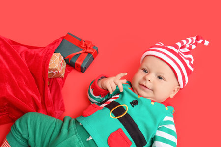Cute little baby in elf's costume and with Christmas gifts on color backgroundの写真素材