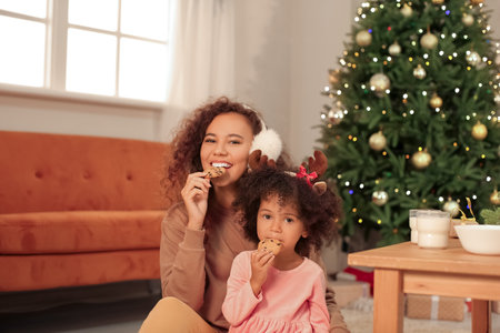 Cute African-American girl and her mother eating cookies at home on Christmas eve
