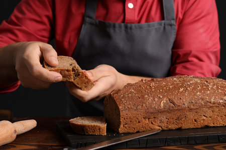Male baker with slice of rye bread at table on dark background, closeup