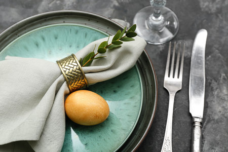 Table setting with Easter egg, cutlery and green branch on gray grunge background