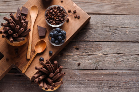 Board with delicious chocolate wafer rolls, coffee beans and blueberries on wooden background