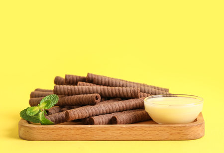 Board with delicious chocolate wafer rolls and condensed milk on yellow backgroundの写真素材