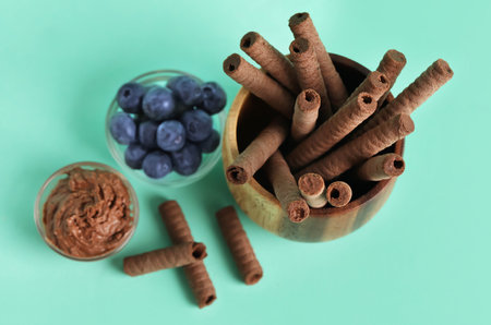 Bowls with delicious chocolate wafer rolls and blueberries on turquoise background