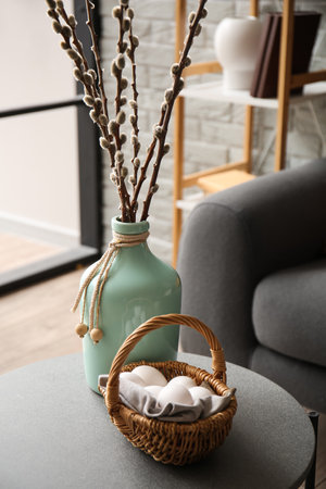 Vase with willow branches and basket of Easter eggs on table in modern living room, closeupの写真素材
