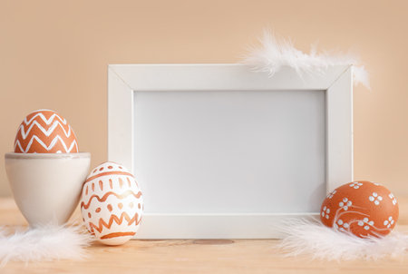 Blank frame, Easter eggs and feathers on wooden table near beige wallの写真素材