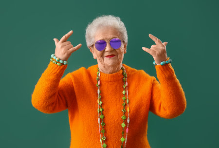 Senior woman with bright accessories showing i love you gesture on green background