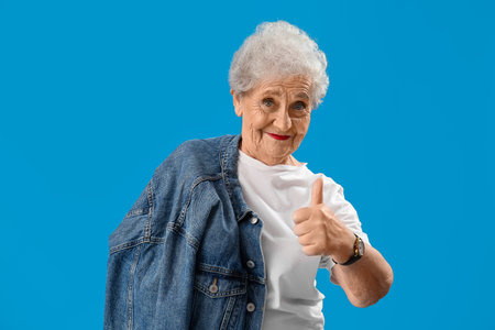 Senior woman with denim jacket showing thumb-up on blue background