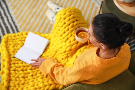 Beautiful young woman with warm plaid reading book and drinking tea at homeの写真素材