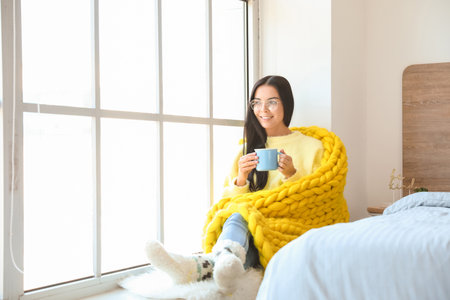 Beautiful young woman wrapped in warm plaid drinking tea near windowの写真素材