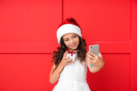 Cute little girl in Santa hat taking selfie on color background
