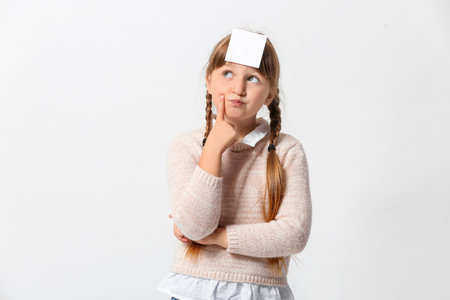 Thoughtful little girl with blank note paper on her forehead against light backgroundの写真素材