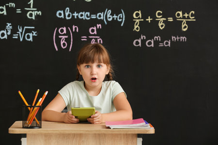 Cute pupil with calculator sitting at desk during lesson in classroom