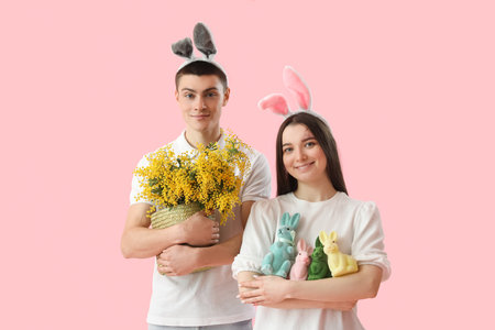 Happy young couple in bunny ears with Easter rabbits and mimosa flowers on pink background
