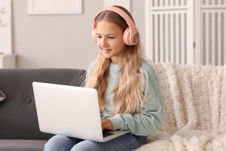 Little girl in headphones using laptop on sofa at homeの写真素材