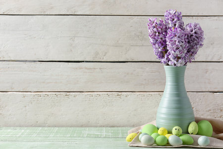 Vase with beautiful hyacinth flowers and painted Easter eggs on green table near white wooden wallの写真素材