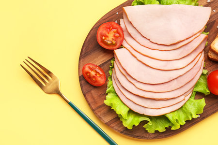 Board with slices of tasty ham, lettuce and tomatoes on yellow backgroundの写真素材