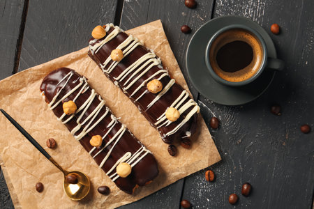 Parchment with delicious chocolate eclairs, hazelnuts and cup of coffee on black wooden background