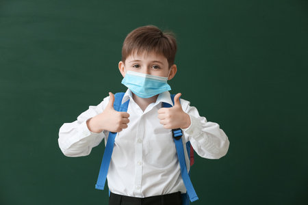 Little schoolboy in medical mask showing thumb-up on color background. Coronavirus epidemic