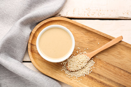 Board with bowl of tasty tahini and sesame seeds on light wooden background, closeupの写真素材