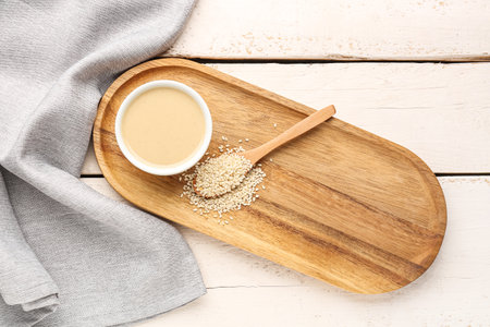 Board with bowl of tasty tahini and sesame seeds on light wooden backgroundの写真素材