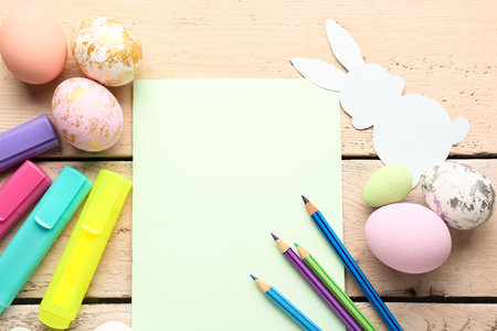 Blank card with Easter eggs, paper rabbit, markers and pencils on beige wooden backgroundの写真素材