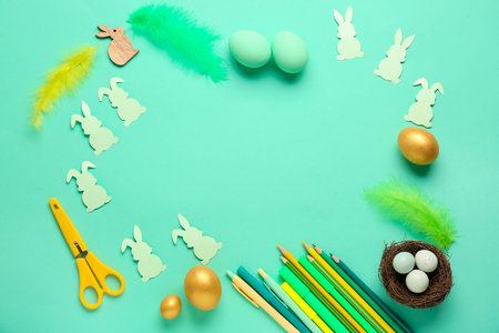 Frame made of Easter eggs, paper rabbits, feathers and pencils on green background