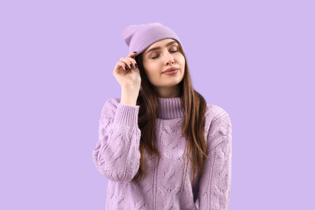 Pretty young woman in knitted sweater and hat on lilac backgroundの写真素材